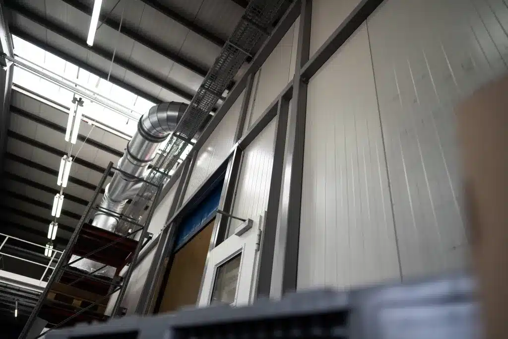 Foto aus Bodenhöhe nach oben gerichtet aufgenommen. Man sieht Metall-Abzugrohre, Industrielampen und eine Hallen-Wand mit Schallschutz