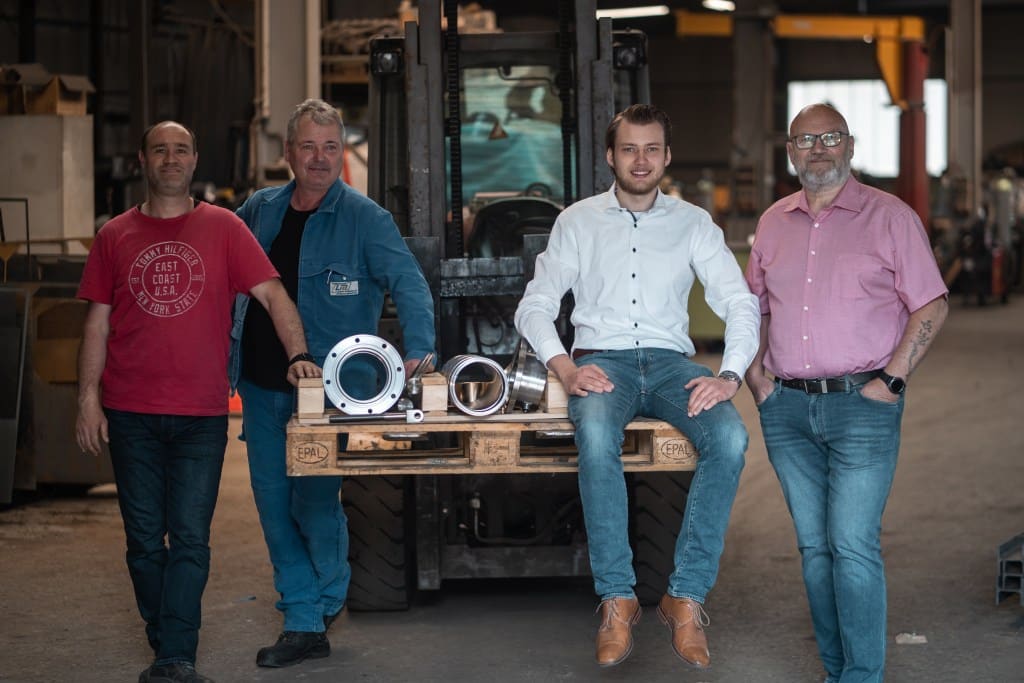 Ein Gruppen Bild von vier Männern vor einem Gabelstapler. Der Gabelstapler hat eine Euro-Palette geladen und der zweite Herr von rechts sitzt auf dieser Europalette.