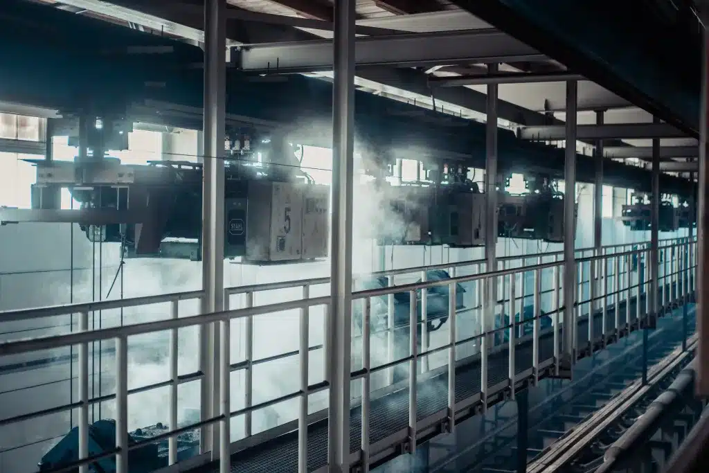 Man sieht einen hoch gelegenen Laufsteg aus Stahl in einer Industriehalle sowie mehrere unter der Decke montierte Kranbahnen. Dies sind gefertigte Lastaufnahmemittel/Lasthebemittel.