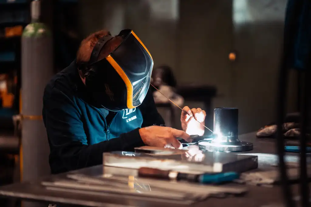 Stahlbau 9 Ein Mann schweißt eine Röhre an eine runde Scheibe mit Löchern fest. Er hält das Schweißmaterial in der linken Hand und das Schweißgerät in der rechten. Er trägt eine Schweißermaske in den Farben von Metallbau Lassner