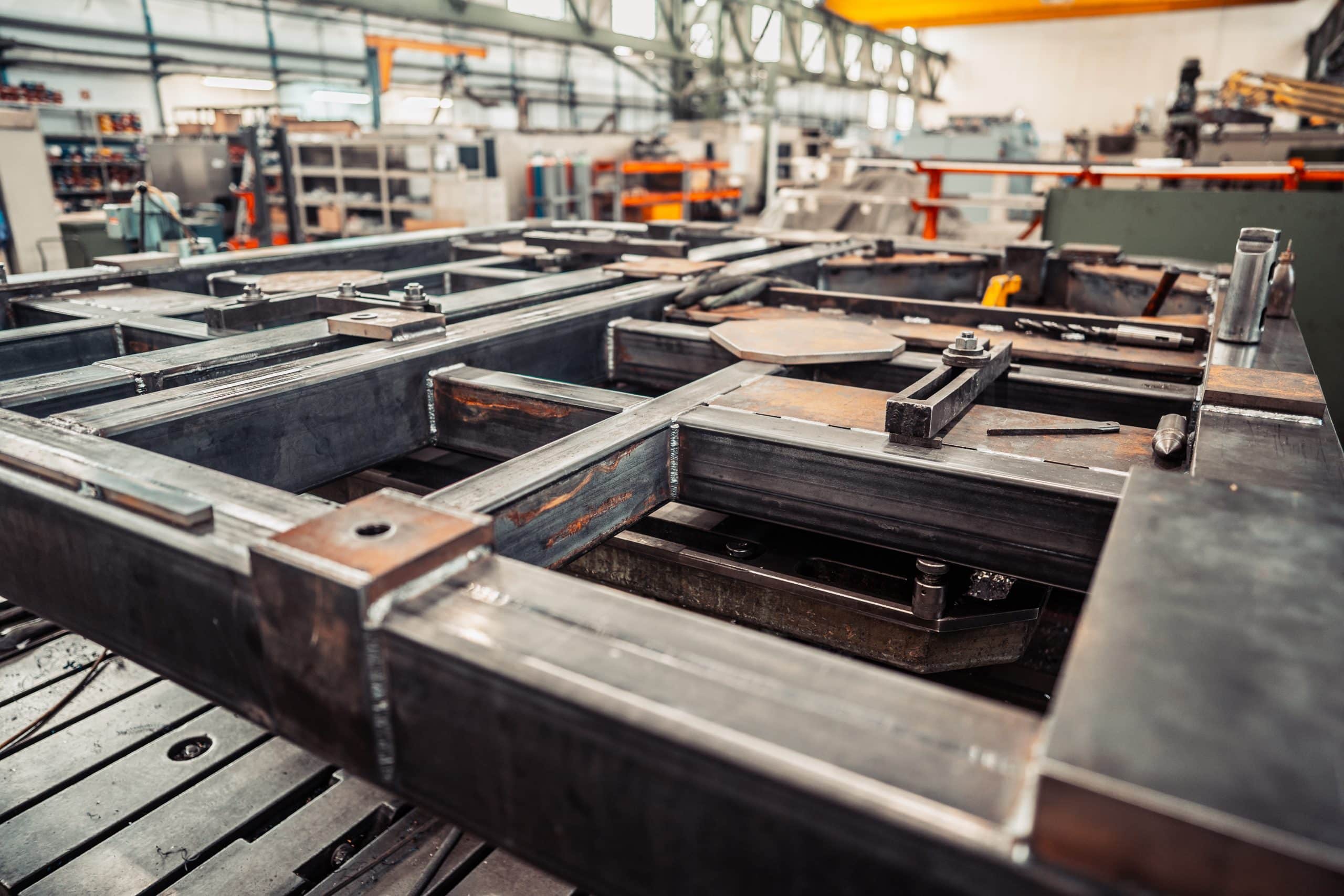 Man sieht ein Stahlgrundgerüst eines Verfahrwagens mit vielen Querstreben, die fachmännisch angeschweißt wurden. Der Hintergrund zeigt eine große Industriehalle in der stahlverarbeitenden Industrie