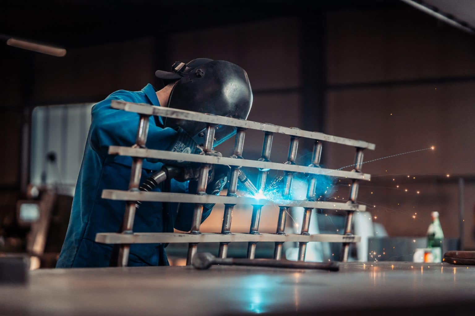 Ein Mann schweißt eine Gitterähnliche Struktur. Es gibt 5 Querstreben und 7 vertikal angeschweißte Röhren. Er trägt eine schwarze Schweißermaske und eine schwarze nach hinten gerichtete Cappie sowie einen Blaumann