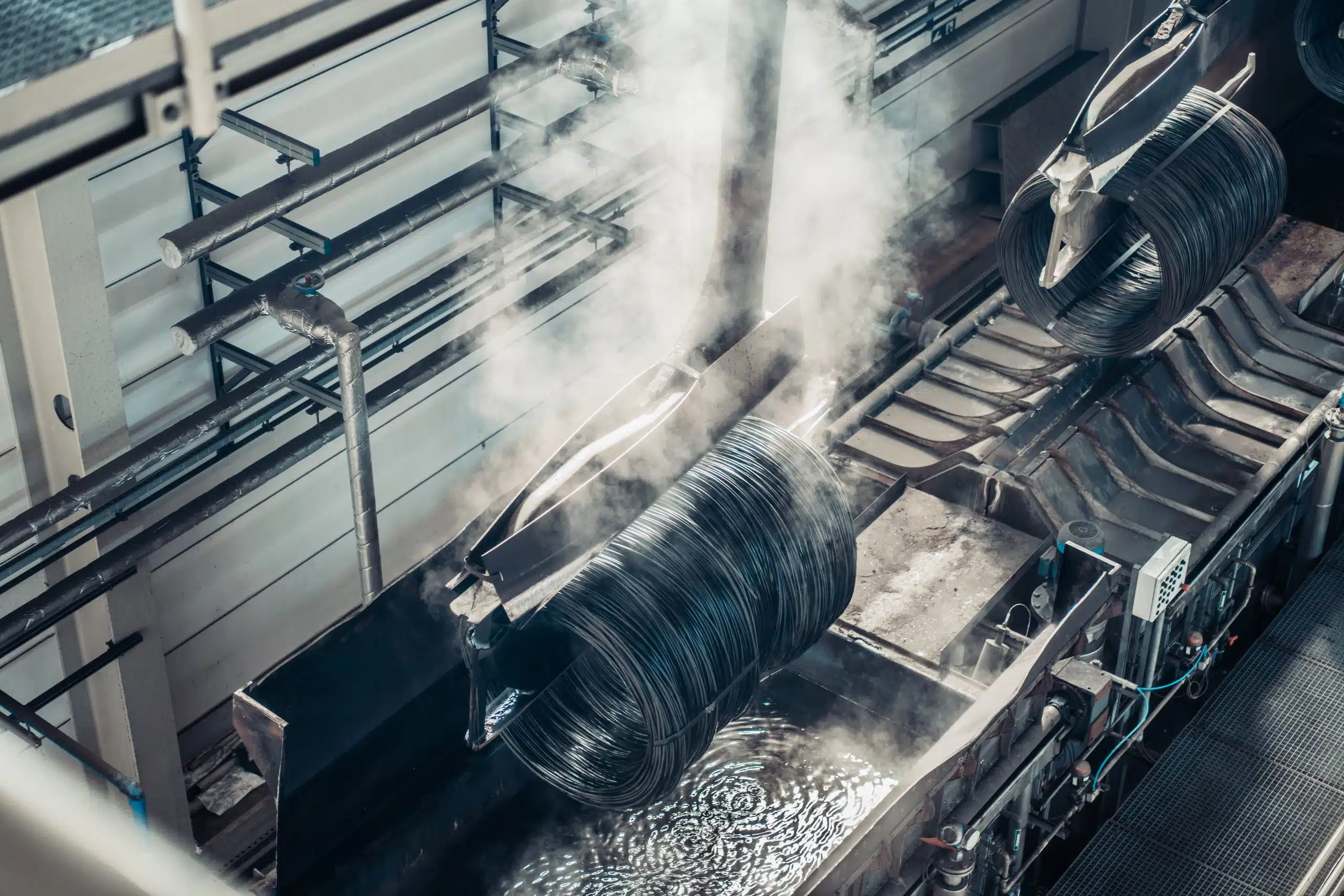 Drahtcoils werden mit einem Kran transportiert und nach und nach in Wasserbecken eingetaucht. Das kürzlich eingetauchte Drahtcoil dampft