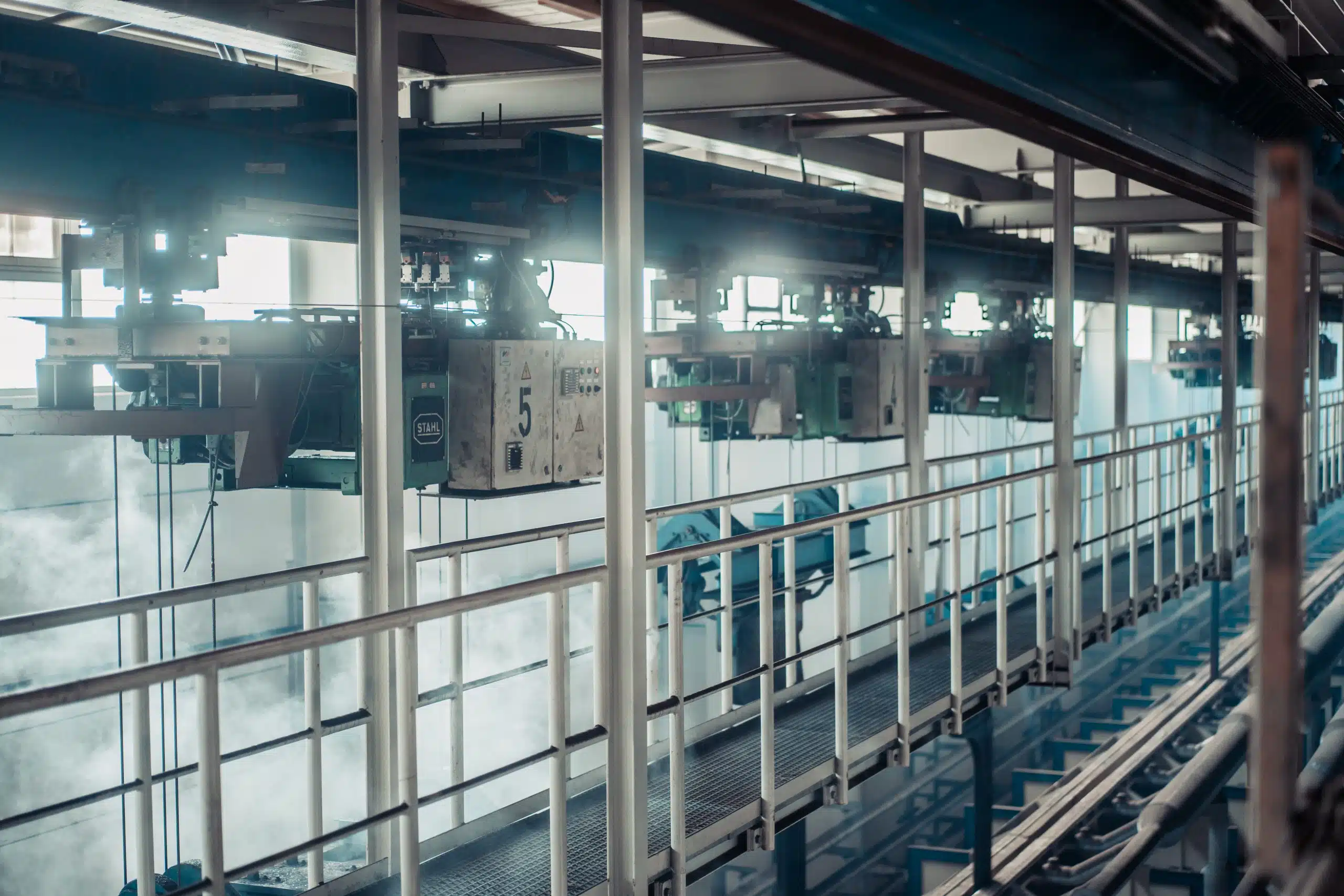 Man sieht einen hoch gelegenen Laufsteg aus Stahl in einer Industriehalle sowie mehrere unter der Decke montierte Kranbahnen. Dies sind gefertigte Lastaufnahmemittel/Lasthebemittel.