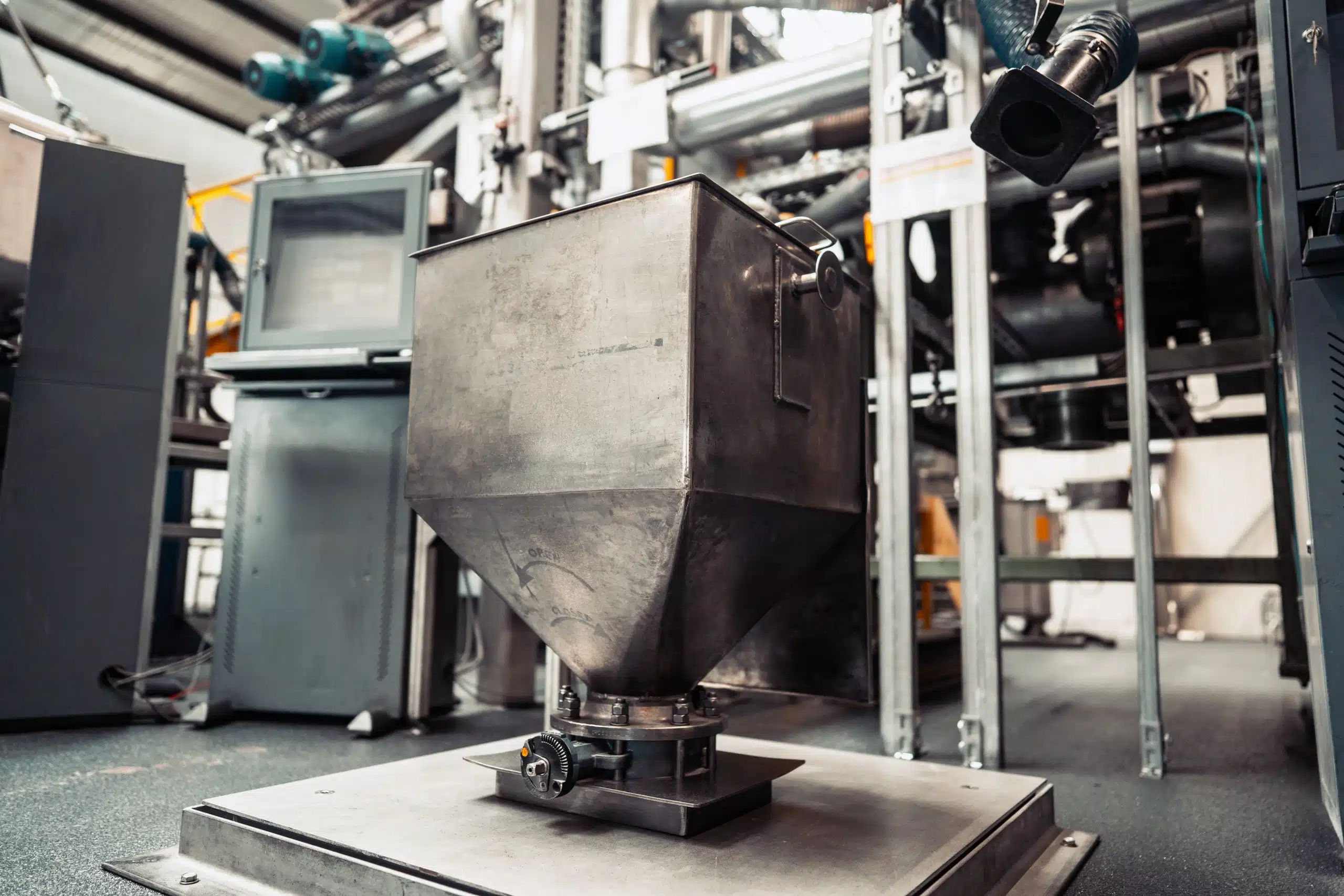 Ein großer Stahltrichter mit einem Gewinde zum Öffnen und Schließen der Lucke am unteren Ende. Der Behälter ist an eine große, am Boden montierte Stahlplatte montiert.